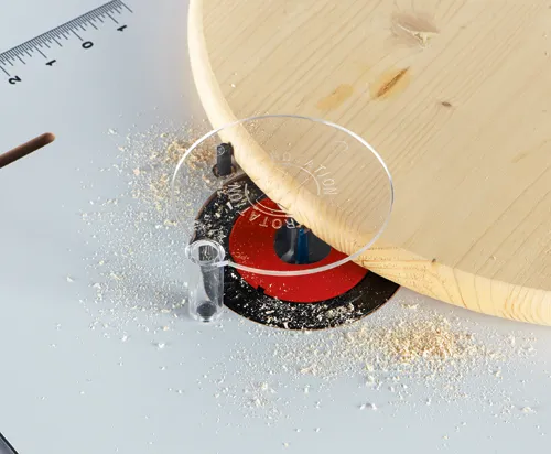 Una pieza de plástico transparente con el texto "ROTATION", colocada en una sierra circular, cortando una tabla de madera, con serrín y una regla.