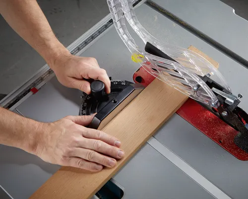Persona utilizando una sierra de mesa, guiando una pieza de madera hacia la hoja. Una protección de seguridad está colocada.