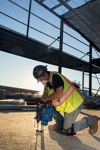 Trabajador de la construcción utilizando un taladro percutor Bosch. El trabajador está arrodillado, usando equipo de seguridad, trabajando bajo una estructura de metal y vidrio.