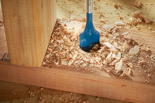 Broca azul perforando un agujero en un trozo de madera, creando virutas de madera.