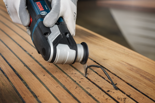 Herramienta multifunción Bosch cortando un cable negro sobre una superficie de madera. La herramienta es sujetada por una persona que lleva guantes.