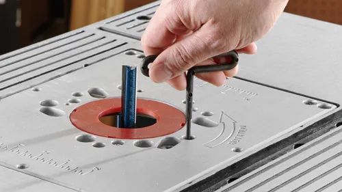 Aquí está la traducción:

Mano usando una llave Allen para ajustar la altura de una fresa en una mesa de metal. La palabra "ROTACIÓN" es visible.