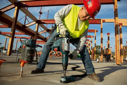 Una persona que lleva equipo de seguridad utiliza un Boschhammer para taladrar hormigón, con una aspiradora Bosch visible en el fondo.