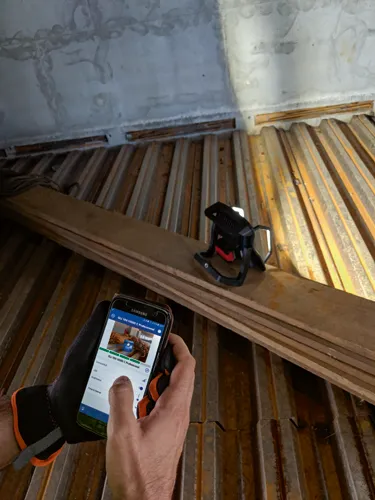 Una persona sosteniendo un teléfono Samsung que muestra "GLI 18V-10000 C Professional" en su pantalla, utilizando la aplicación para ajustar el brillo de una lámpara de trabajo. También es visible una lámpara de trabajo Bosch GLI 18V-10000 C Professional.