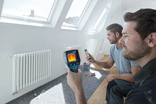 Dos personas observan un radiador caliente utilizando una cámara termográfica Bosch y un smartphone, interior de una habitación con tragaluces.