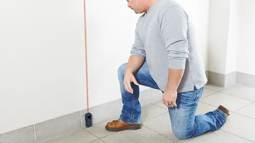 Persona arrodillada cerca de una pared mientras utiliza un medidor láser rojo; la herramienta muestra "9" y "10".