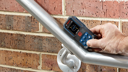 Bosch BLAZE laser measure displays -32.0° en pantalla. La mano sostiene el dispositivo contra una barandilla metálica, frente a una pared de ladrillo.