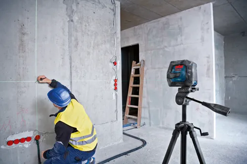 Bosch GLL300-40G láser de líneas, montado sobre trípode, proyectando líneas verdes; trabajador de la construcción con casco azul marca pared.
