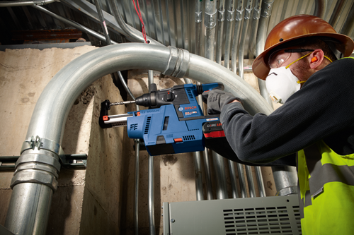 Bosch GBH18V-26, un martillo rotativo azul y negro, sostenido por una persona que lleva equipo de seguridad, taladrando hormigón junto a tuberías metálicas.