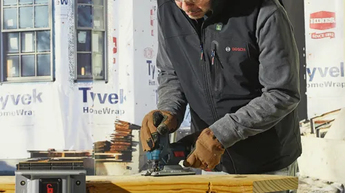 Persona que lleva una chaqueta negra con el logotipo "BOSCH", utilizando una sierra de calar, con "Tyvek" visible en el fondo.