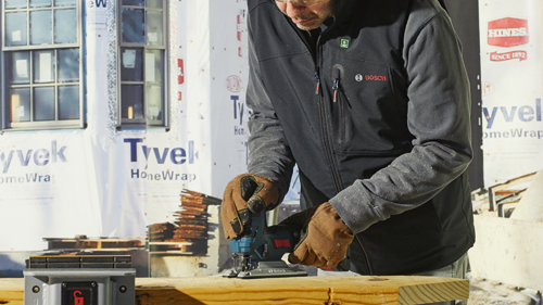 Persona que lleva una chaqueta negra con el logotipo "BOSCH", utilizando una sierra de calar, con "Tyvek" visible en el fondo.