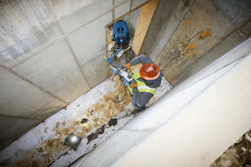 Persona que lleva casco y chaleco de seguridad utiliza una herramienta eléctrica en una pared de hormigón; una aspiradora está cerca.