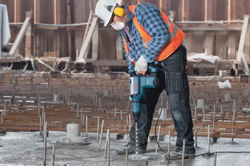 Persona utilizando un martillo demoledor Bosch RH850VC en una obra, con casco de seguridad, protección auditiva y chaleco de seguridad naranja.