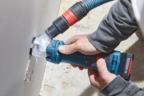 Herramienta Bosch siendo utilizada en una pared blanca, con una manguera de aspiración conectada y la palabra "BOSCH" visible en la batería.