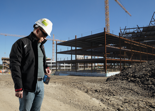 Hombre con casco blanco y gafas de sol en una obra, sosteniendo una batería Bosch CORE18V; son visibles grúas de construcción y la estructura de un edificio en construcción.