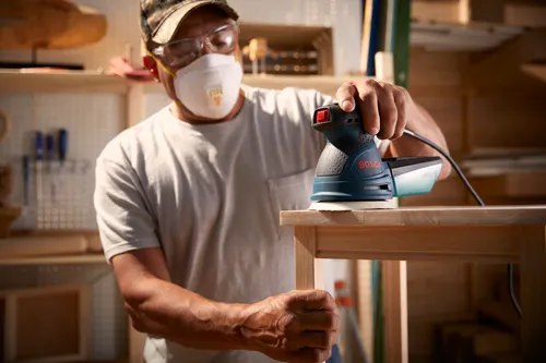 Hombre con gafas de seguridad y mascarilla lijando una mesa de madera con una lijadora Bosch.
