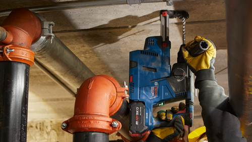 Bosch GBH18V-24C BULLDOG, sostenido por persona con guantes amarillos, taladrando un agujero en un techo. Tuberías rojas y negras visibles.