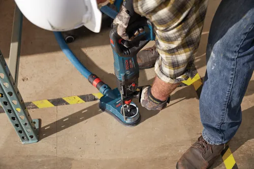 Persona utilizando un martillo rotativo Bosch Bulldog GBH18V-26D, con un accesorio de aspiración de polvo DC200, vistiendo una camisa a cuadros y vaqueros.
