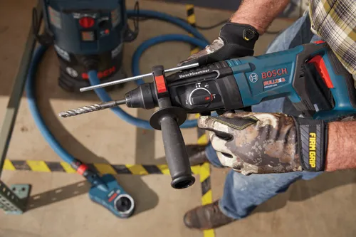 Bosch GBH18V-26D, martillo rotativo Bulldog, con SDS, sostenido por persona que lleva guantes. Una aspiradora azul está en el fondo.