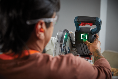 Una persona que lleva gafas de seguridad y protección auditiva opera una sierra eléctrica con una pantalla "TURBO" que muestra la velocidad como "3", un ajuste "ECO" y el nivel de la batería.