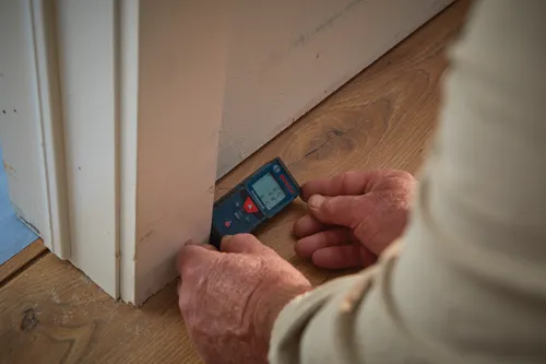 Medidor de distancia láser Bosch Professional, sostenido por una persona sobre un suelo de madera cerca de un marco de puerta; la pantalla muestra mediciones.