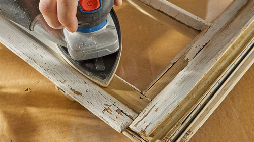 Persona utilizando una lijadora en un marco de ventana de madera pintado de blanco y antiguo.