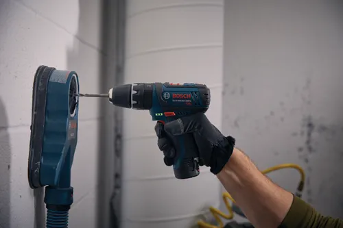 Persona que lleva un guante sostiene un taladro Bosch de 12V de iones de litio frente a una pared, con una broca insertada en una campana de extracción de polvo Bosch HDC200 acoplada.