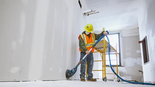 Persona lijando una pared con una herramienta en una zona de construcción. Lleva un casco de seguridad amarillo y un chaleco de seguridad naranja.