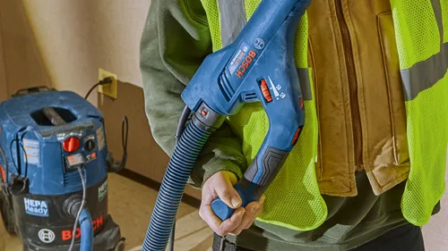 Una persona con un chaleco de seguridad sostiene una lijadora Bosch GTB55-85, cerca de otro aparato Bosch, HEPA Ready.