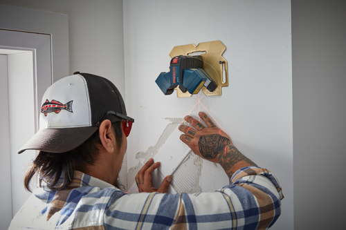 Una persona instala una baldosa, utilizando un nivel láser Bosch GTL3. El nivel láser está montado en una pared y proyecta líneas rojas. La persona lleva una camisa a cuadros y una gorra con un gráfico de pez.