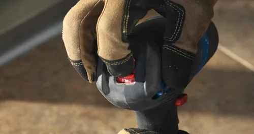 Mano enguantada presionando un botón rojo en una herramienta negra y azul, con "930°F" impreso en ella.