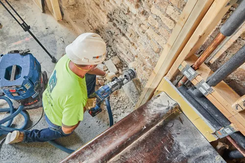 Una persona que lleva un casco, gafas de seguridad, guantes y una camisa verde neón utiliza un taladro percutor Bosch CORE18V junto a una aspiradora Bosch HEPA Ready. La persona está taladrando una pared de ladrillo. Adyacente hay tuberías y estructura expuestas.