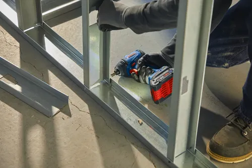 Persona con guantes utilizando un atornillador de impacto Bosch GDX18V-1800C en un marco metálico, batería CORE18V 4.0 Ah visible.