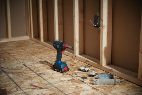 Destornillador de impacto inalámbrico Bosch sobre suelo de contrachapado cerca de estructura, caja eléctrica y enchufe.