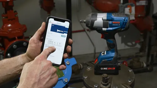 Llave de impacto a batería Bosch, GSR18V-740C, y un smartphone mostrando la interfaz de control para configurar y guardar los ajustes de liberación de pernos.