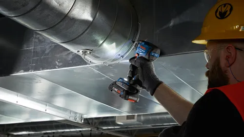 Una persona que lleva un casco amarillo y gafas de seguridad, instalando un conducto de aire con un taladro inalámbrico Bosch GSB18V-180C.