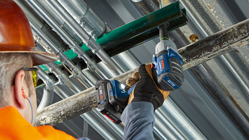 Persona que lleva casco naranja y gafas de seguridad utilizando una llave de impacto inalámbrica Bosch, con texto visible 'Bosch' y 'BRUSHLESS MOTOR', instalando sobre tuberías.