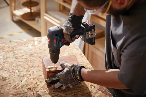 Una persona que lleva guantes y gafas de seguridad utilizando un taladro atornillador a batería Bosch GSB18V-490 para perforar un agujero en una caja de madera.