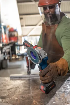 Una persona que lleva equipo de seguridad sostiene una amoladora angular Bosch CORE18V. La amoladora está en uso y la marca "BOSCH" es visible en su empuñadura.