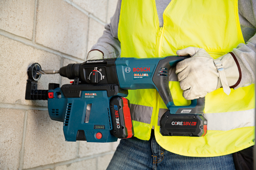 Bosch BULLDOG GBH18V-26D martillo rotativo inalámbrico con baterías CORE18V, sostenido por una persona que lleva guantes de trabajo y un chaleco amarillo mientras taladra en una pared de bloques de hormigón.