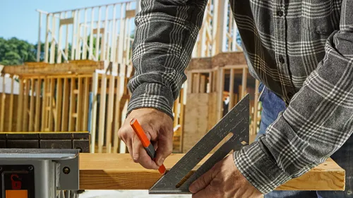 Persona utilizando una escuadra de velocidad para marcar una tabla de madera, construcción al fondo.