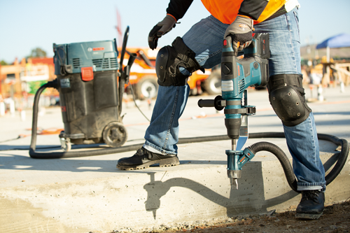 Bosch GBH18V-36C, una persona utilizando un taladro y una aspiradora. La persona lleva equipo de seguridad, incluyendo guantes y rodilleras. Se ve una aspiradora HEPA.