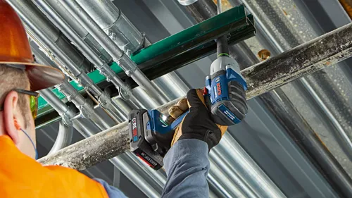 Una persona que lleva gafas de seguridad y un casco naranja utiliza una llave de impacto Bosch con 'BRUSHLESS MOTOR' para trabajar en tuberías metálicas, con una batería 'CORE18V' acoplada.