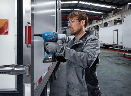 Hombre con gafas utilizando una remachadora ciega Bosch GRG18V-3600C con batería CORE18V. La herramienta está acoplada a una puerta de vehículo.