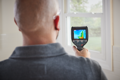 Una persona con una cámara termográfica apuntando hacia una ventana, la pantalla mostrando lecturas de temperatura.