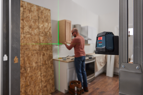 Bosch Professional GLL50-20G nivel proyectando una línea láser verde, una persona instalando un mueble de cocina en el fondo.