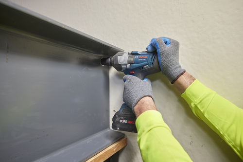 Herramienta eléctrica Bosch GDS18V-1180C TURBO BRUSHLESS utilizada para perforar una viga metálica gris, persona con guantes grises y camisa amarilla de manga larga sujetando la herramienta.