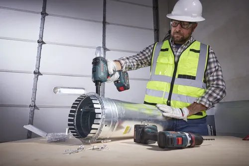 Una persona que lleva gafas de seguridad, un casco blanco y un chaleco de seguridad reflectante utiliza una remachadora inalámbrica Bosch GFR18V-21 en un tubo de metal. Otro taladro Bosch y una batería adicional están sobre una superficie de madera.