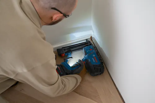 Una persona con gafas y guantes de trabajo, utilizando una clavadora inalámbrica Bosch GNH18V-18M en una esquina de una pared.