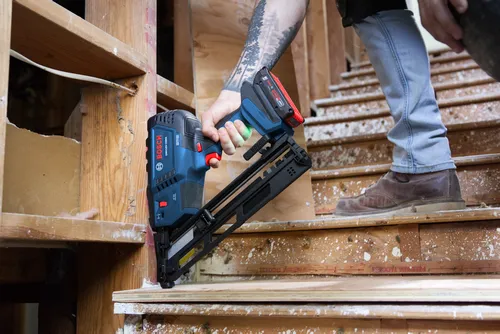 Claro, aqui está a tradução:

Clavadora inalámbrica Bosch GNH18V-15MA sostenida por una persona con un tatuaje, de pie cerca de unas escaleras de madera.